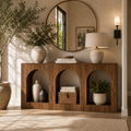 Arched wood console table styled beneath a round mirror with vases, books, a basket, greenery, and a lamp.