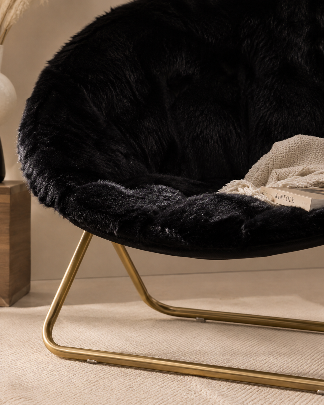 A close-up of a modern round accent chair with a black faux fur cushion and a curved gold metal base, styled with a beige knit blanket and a book in a warm indoor setting.