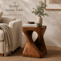 The Sculpt wood accent table styled with books, a bowl, and leafy branches beside a neutral sofa.